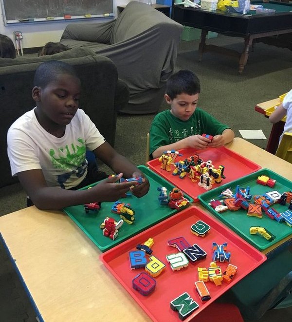 Two preschoolers sorting letter blocks on trays at a classroom table